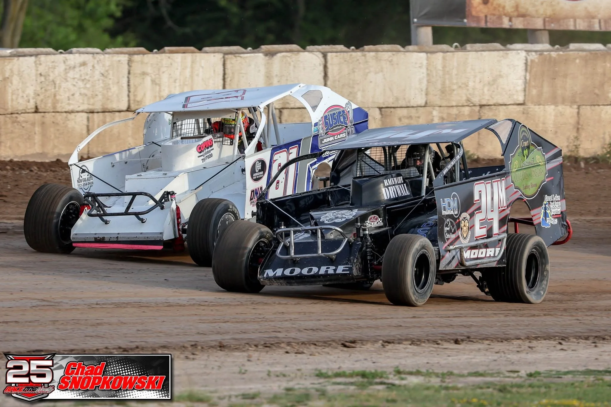 Two modified race cars competing on a dirt track during a race, with a concrete barrier in the background.