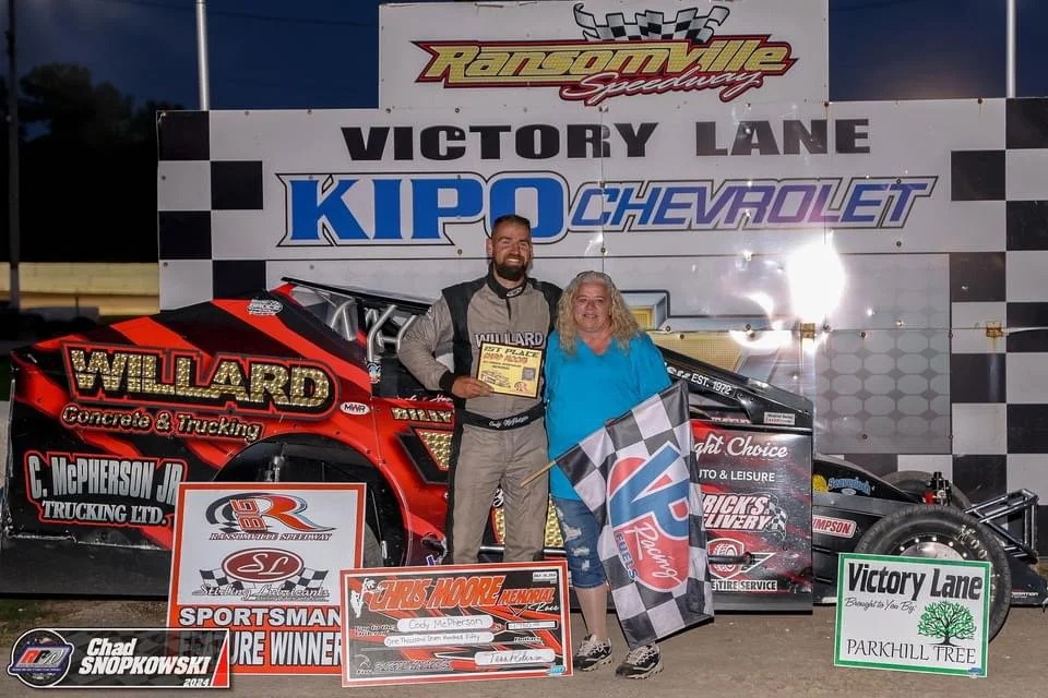 A race car driver and a woman standing on a race track victory lane, holding a checkered flag and a certificate. The background shows a large sign with 
