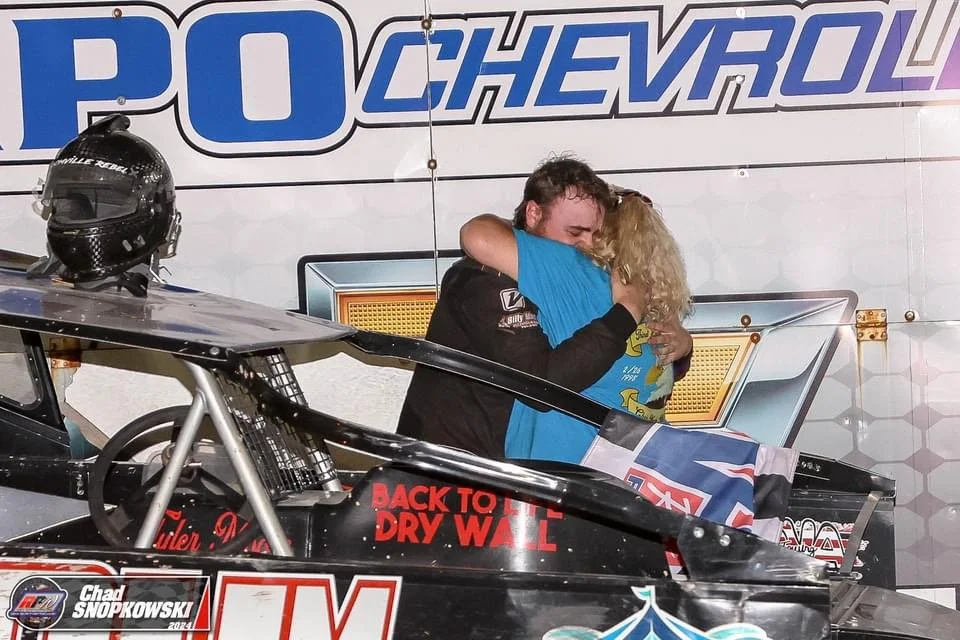 A race car driver hugging a young girl during a celebration, with a race car in the foreground and a race-themed backdrop in the background.