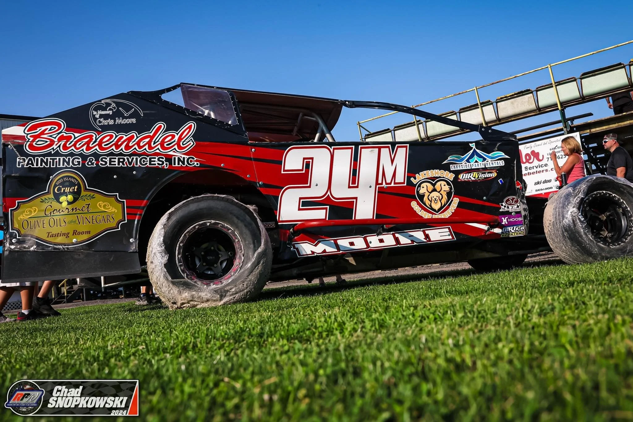 Race car with the number 24, sponsor decals, and a group of people standing nearby on a grassy field under a clear blue sky.