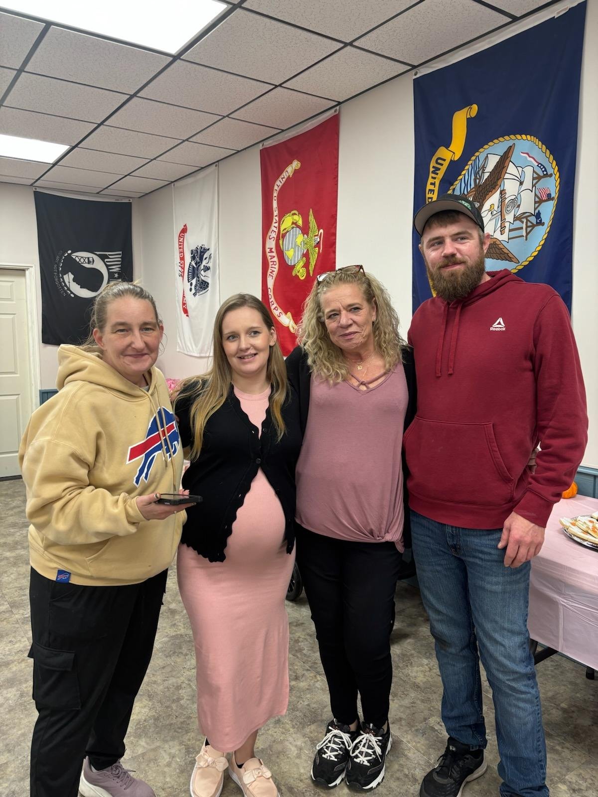 Four people standing together in front of flags, including a woman with a pregnant belly, a woman with curly blonde hair, a woman in a beige hoodie, and a man with a beard in a red hoodie, smiling at the camera.