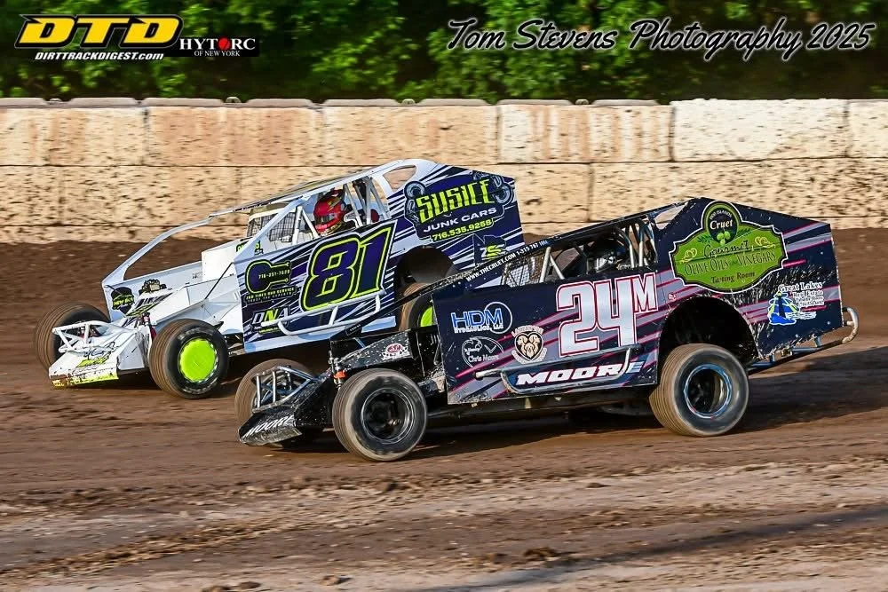 Two dirt track racing cars on a dirt track, with a concrete barrier and green trees in the background. The cars are numbered 81 and 24 and are competing closely during a race.