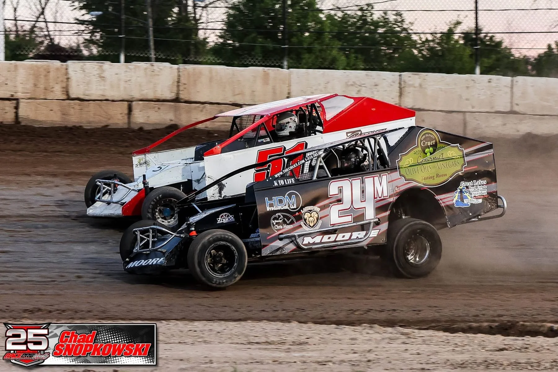 Two dirt track race cars on a dirt track, racing side by side. The car in front is black with red and white accents and the number 24M, while the car behind is white with red and black accents and the number 51. Both cars are kicking up dust as they 