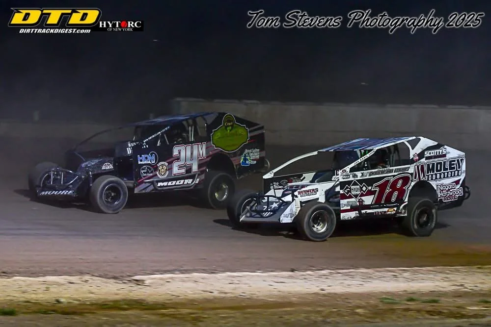 Two race cars on a dirt track at night, number 24 on the left and number 18 on the right, competing closely. The car on the left is black with various logos and the number 24 in large font, while the car on the right is white and black with the numbe