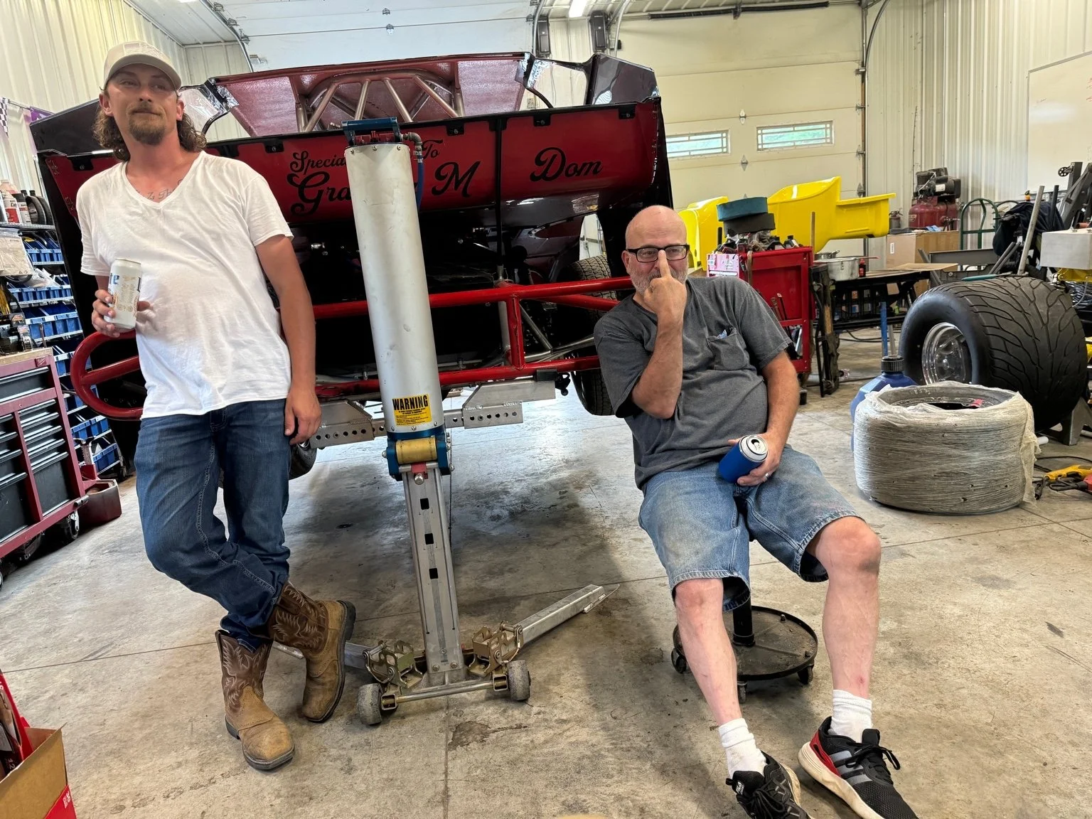 Two men in a workshop with a racecar; one man is standing, wearing a white t-shirt, jeans, and boots, holding a drink; the other man is sitting, wearing a gray t-shirt, shorts, sneakers, and glasses, holding a drink and covering his face with his han