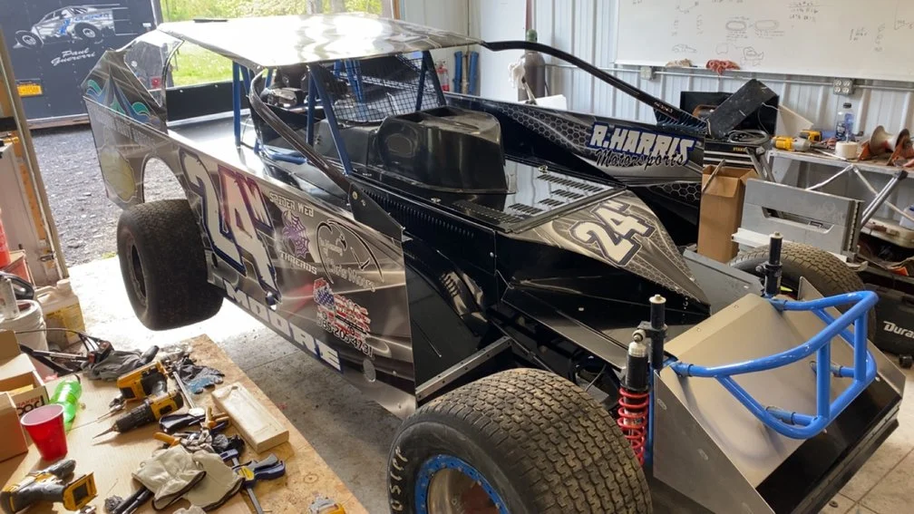 A black racing dirt car with the number 24 and various decals on the side, parked inside a garage with tools and equipment around.