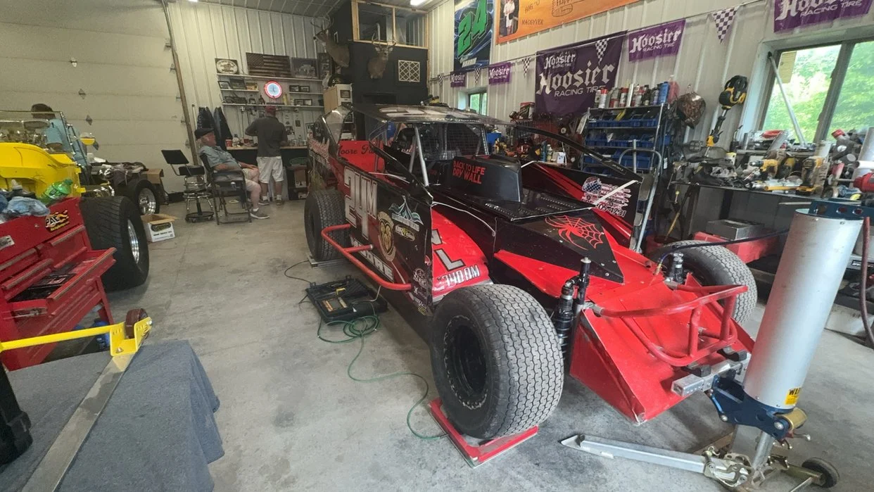 A race car in a garage workshop with tools and equipment around, and a few people working or sitting nearby.