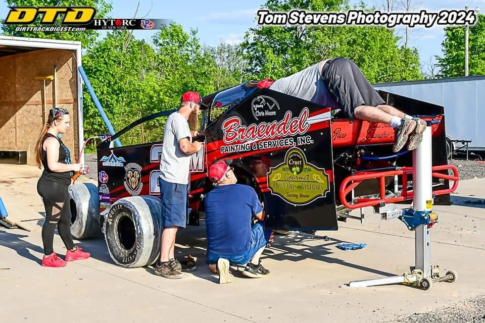 Team members working on a race car, with one person lying inside the car's engine area and two others inspecting or repairing it. The car is black with red and white sponsorship logos, and it has large tires. The scene appears to be at a racing event