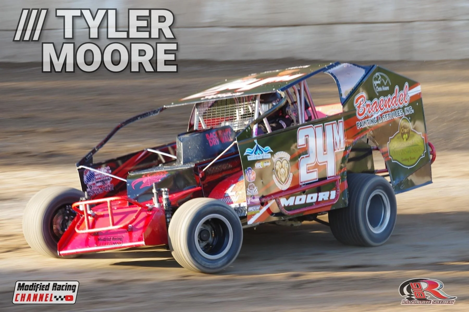 A modified race car with the number 24 on the side, driving on a dirt track at Ransomville Speedway. The car is black with red and gold accents, featuring various sponsor stickers. The driver is inside wearing a helmet, and the background shows a woo