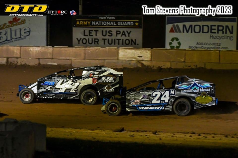 Two race cars on a dirt track at night, with advertisements and signs in the background. One car is black and white with the number 4 on it, and the other is primarily black with blue accents and the number 24.