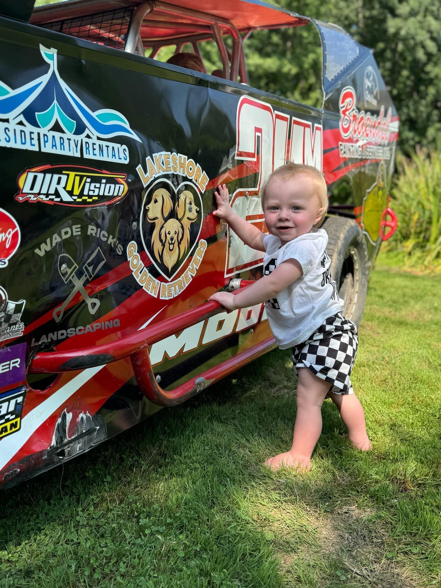 A young child with blonde hair, wearing a white t-shirt and black-and-white checkered shorts, is standing outside on grass, holding onto the side of a race car. The race car is decorated with various sponsor logos and the number 20.