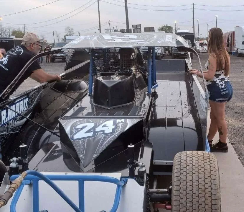 Race car with the number 24, people standing beside it in a parking lot or pit area.
