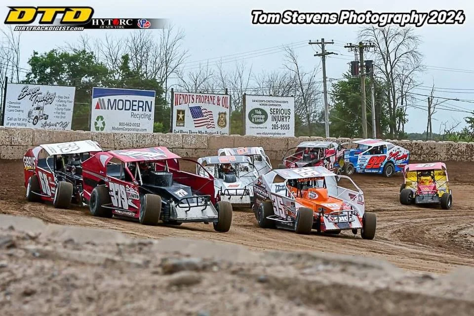 Peat racing cars on dirt track during race, with banners and power lines in background.