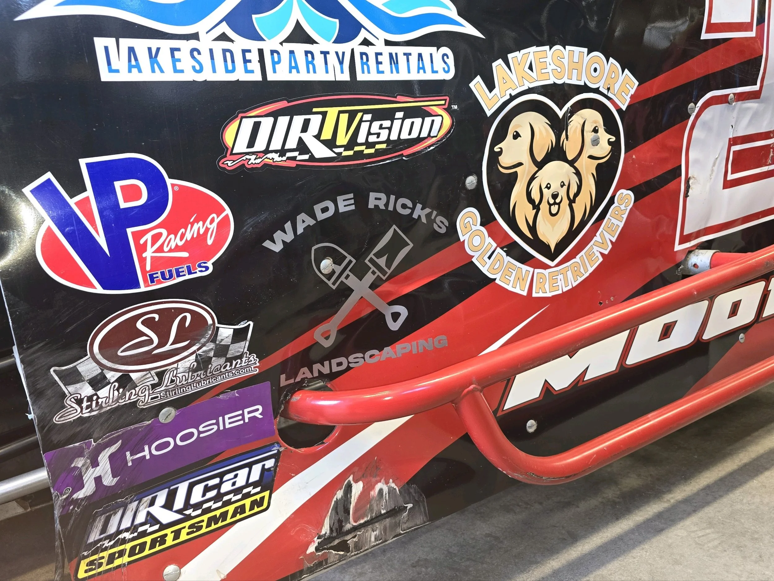 Close-up of a racing vehicle with various sponsor stickers, including Lakeside Party Rentals, DIRTVision, VP Racing Fuels, Sterling Lubricants, Hoosier, and American Racer, along with a sticker for Lakeshore Golden Retrievers.