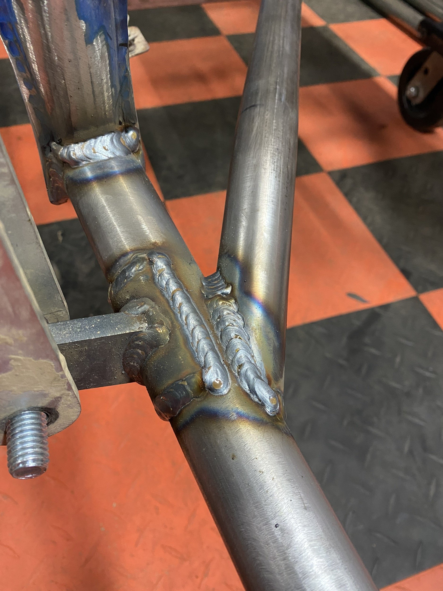 Close-up of a welded metal frame, showing the welds and joints connecting tubular steel components on a checkered floor.