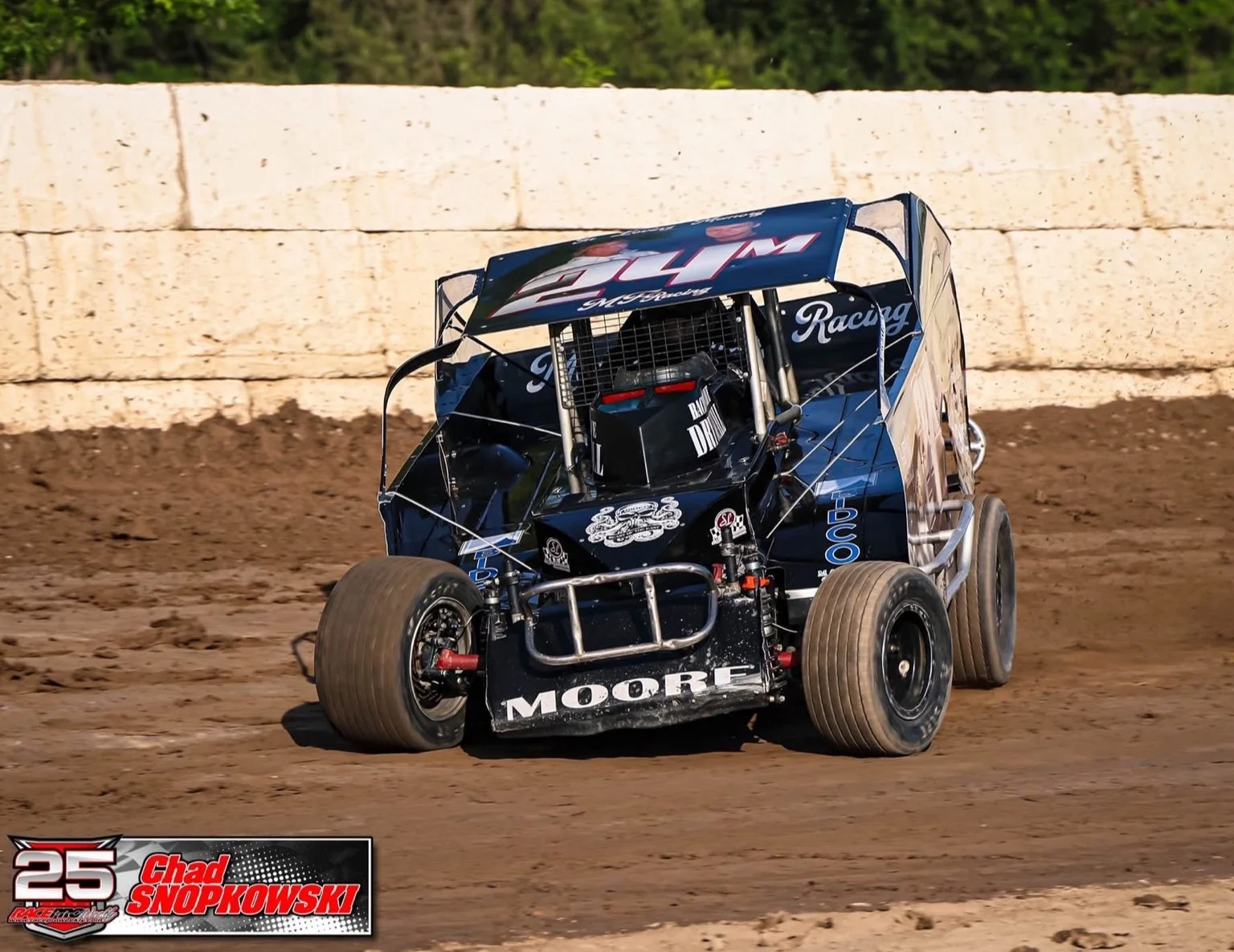 A black and blue DIRT Sportsman racing on a dirt track, with a white wall and trees in the background. The car has the number 24M and sponsor logos, including Moore, displayed on the front and sides.