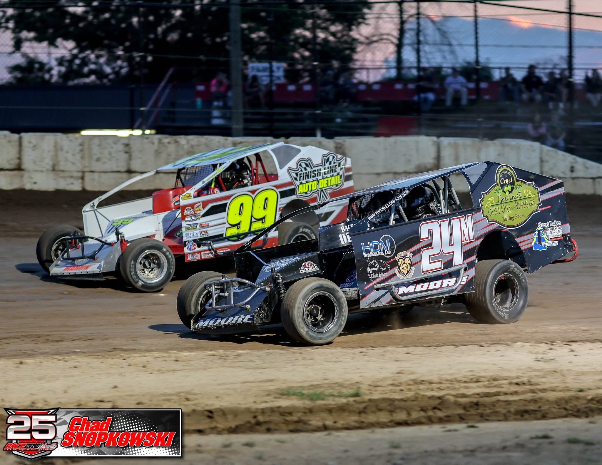 Two dirt track race cars, numbered 99 and 24, competing on a dirt oval track during sunset with spectators in the background.