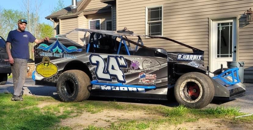 A man standing next to a black and blue race car with the number 24 on it, parked on a grassy area in front of a beige house with a sliding glass door.