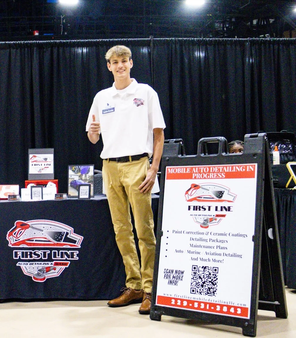 A young man smiling and giving a thumbs up at a booth for First Line Auto Detailing, with a black curtain backdrop and a signboard listing services like paint correction, ceramic coatings, detailing packages, maintenance plans, and auto marine and aviation detailing.