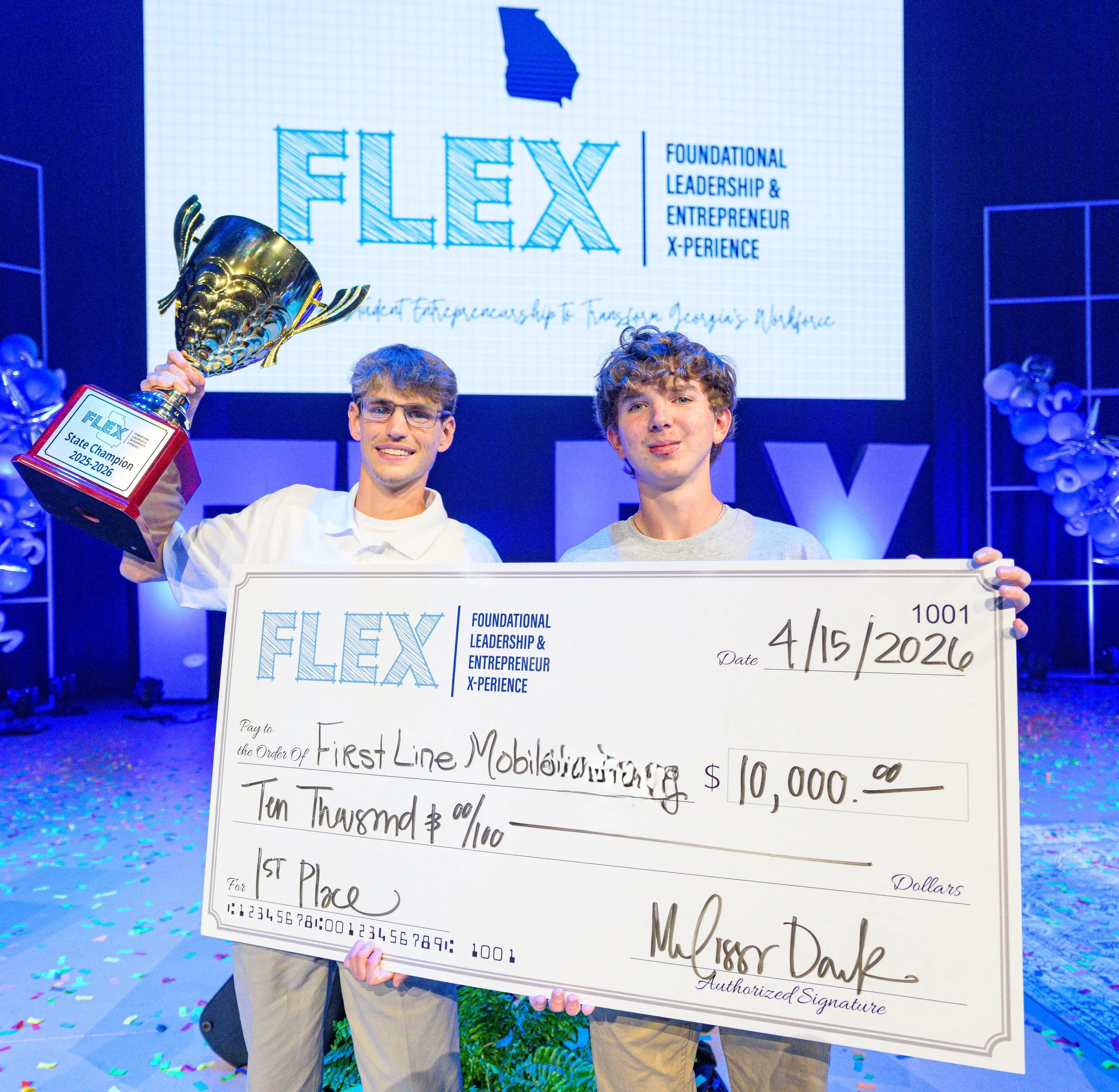 Two young men standing side by side holding a large check and a trophy on stage at an event. The check is made payable to First Line Mobility with a prize amount of $10,000. The background features a screen displaying the event logo 'FLEX' with the words 'Foundational Leadership & Entrepreneur X-Perience'. The stage is decorated with balloons and confetti.