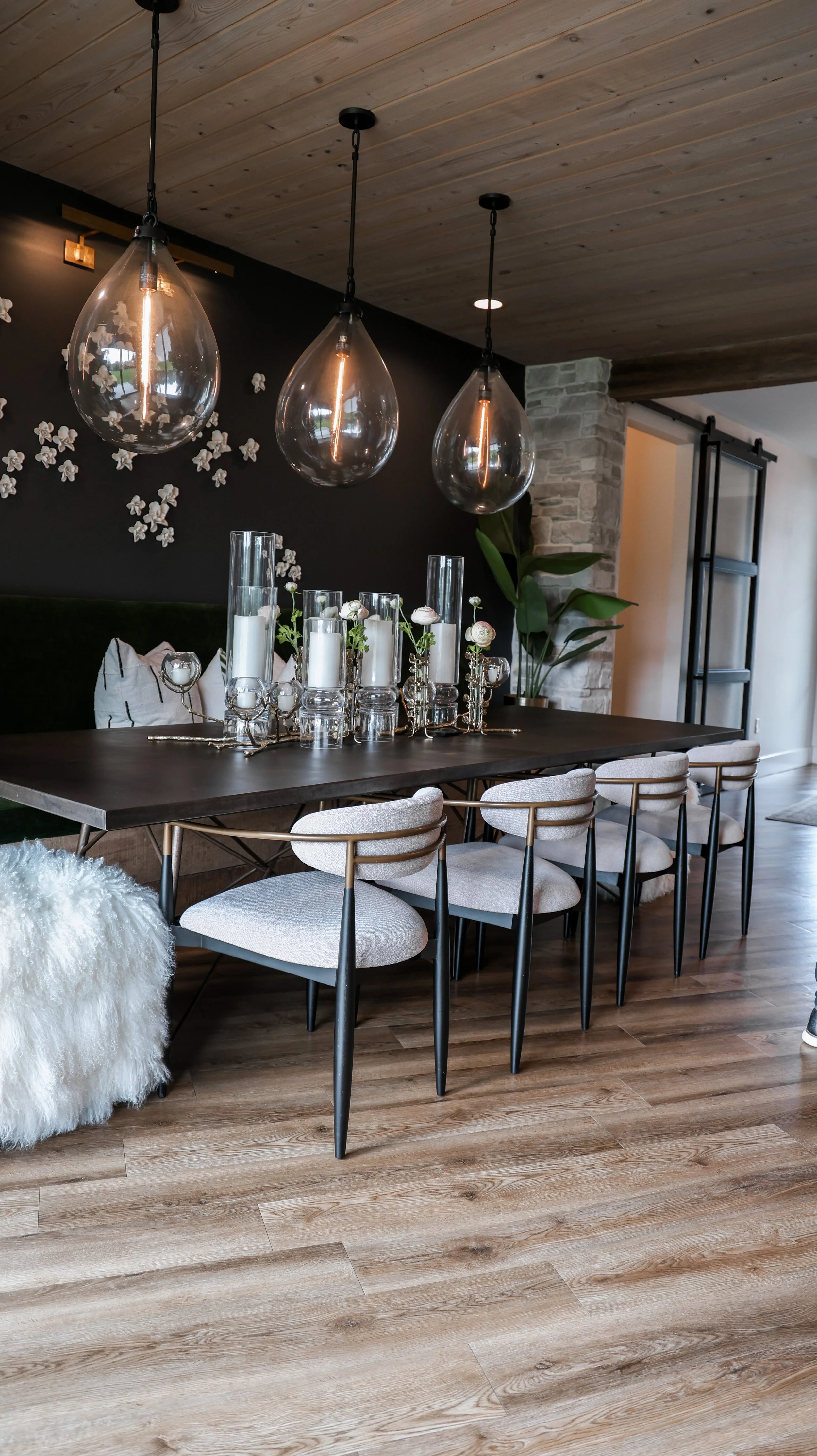 Modern dining room with a dark wood table, eight light-colored upholstered chairs, and pendant lights hanging above. Decor includes white candles, glass vases, and floral accents. Features a black accent wall with decorative floral wall art, wooden c