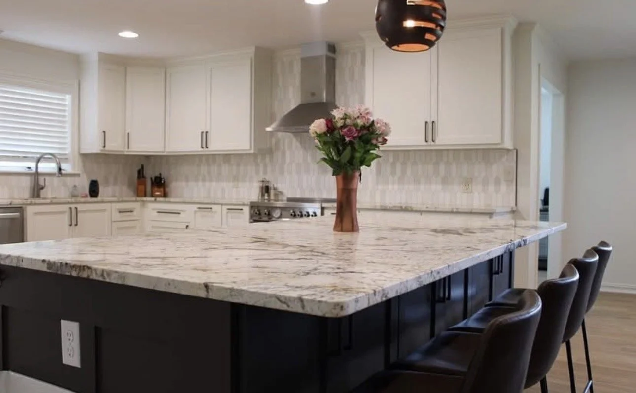 Modern kitchen with white cabinetry, a large marble island with dark base, black barstools, a vase with pink and white flowers, and a hanging black pendant light.
