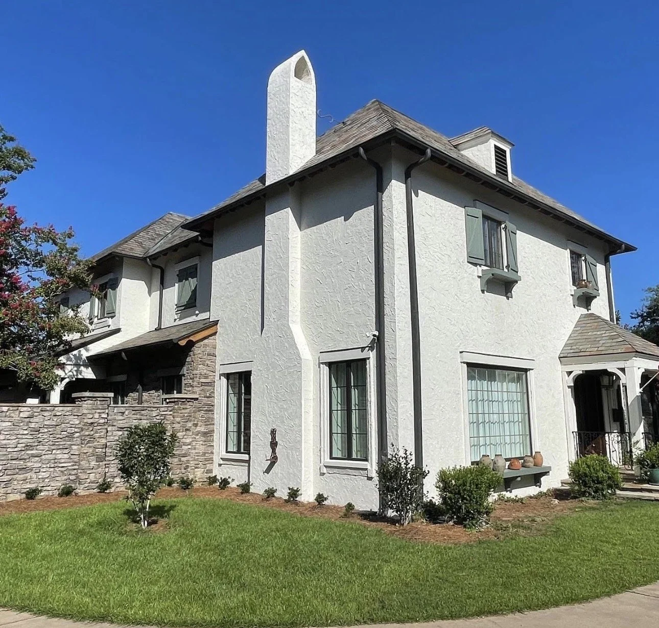 A white two-story house with a stone wall on one side, green lawn, small bushes, and potted plants near the entrance, under a bright blue sky.