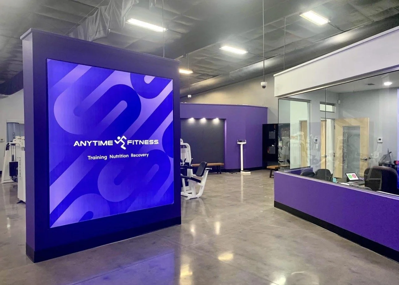 Interior of a fitness center with a large digital screen displaying 'Anytime Fitness, Training Nutrition Recovery'. There are exercise machines, a scale, and a reception area with purple and white accents and glass walls.