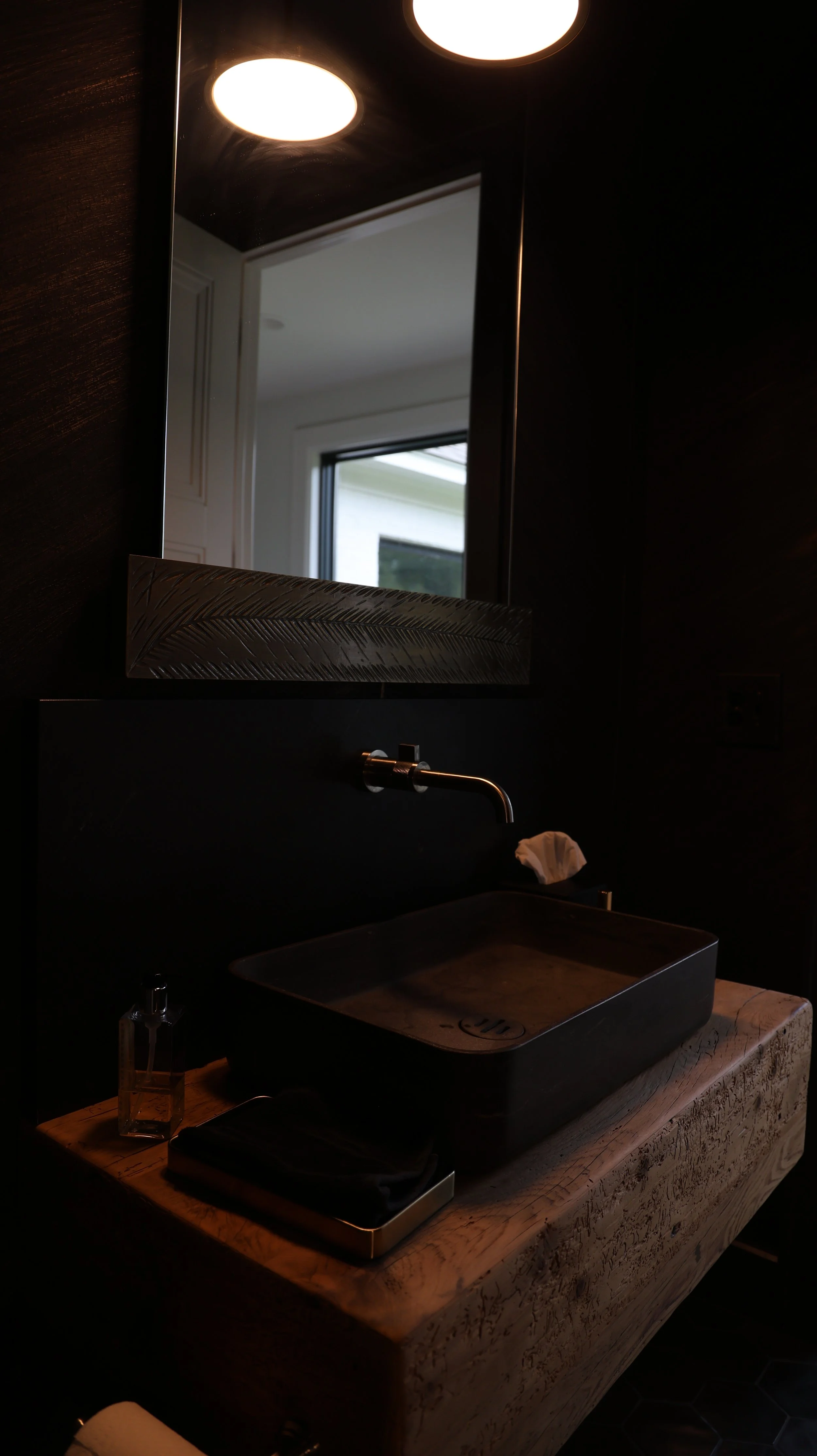A dark bathroom with a large mirror, a black vessel sink on a wooden countertop, and overhead lighting reflecting off the mirror. A wall-mounted faucet and a small soap dispenser are also visible.