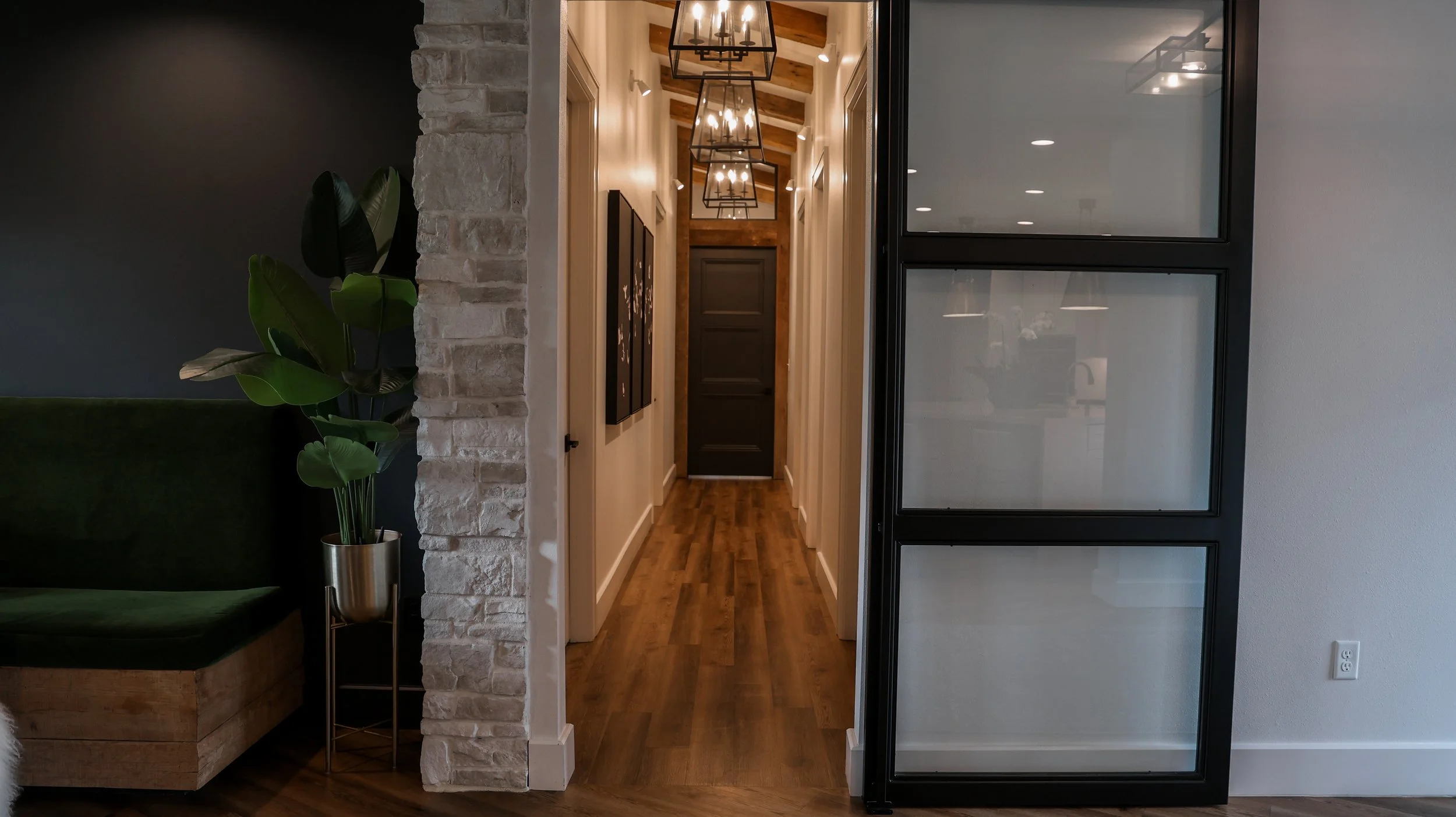 A hallway with wood flooring, white walls, black door at end, framed art on left wall, multiple black metal chandeliers hanging from wooden ceiling beams, seen through a black-framed glass wall on right, with a green sofa and potted plant on the left