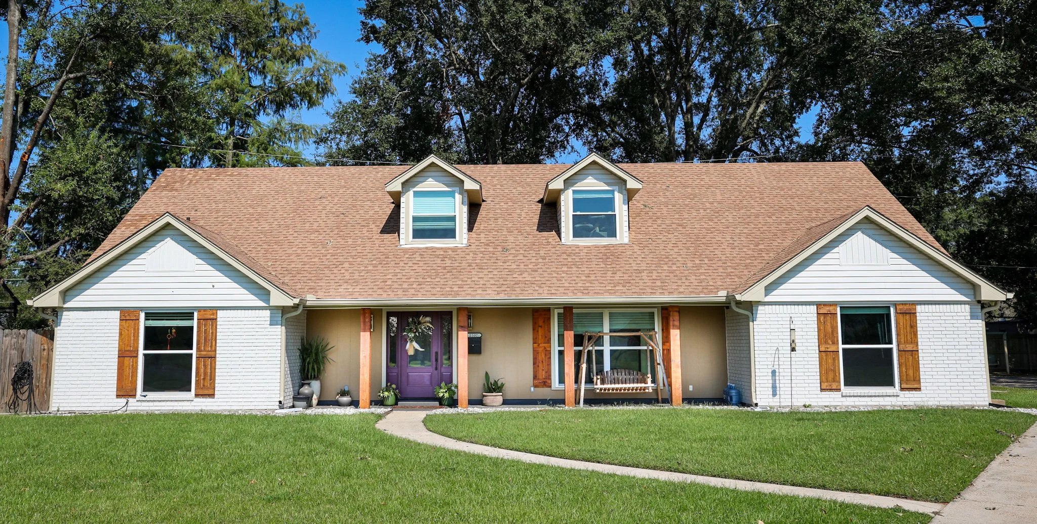 Front view of a house with a brown gable roof, white brick walls, and purple front door. Two dormer windows on the roof with blue shutters, and large windows on each side of the door with wooden shutters. Front yard with green grass, potted plants ne