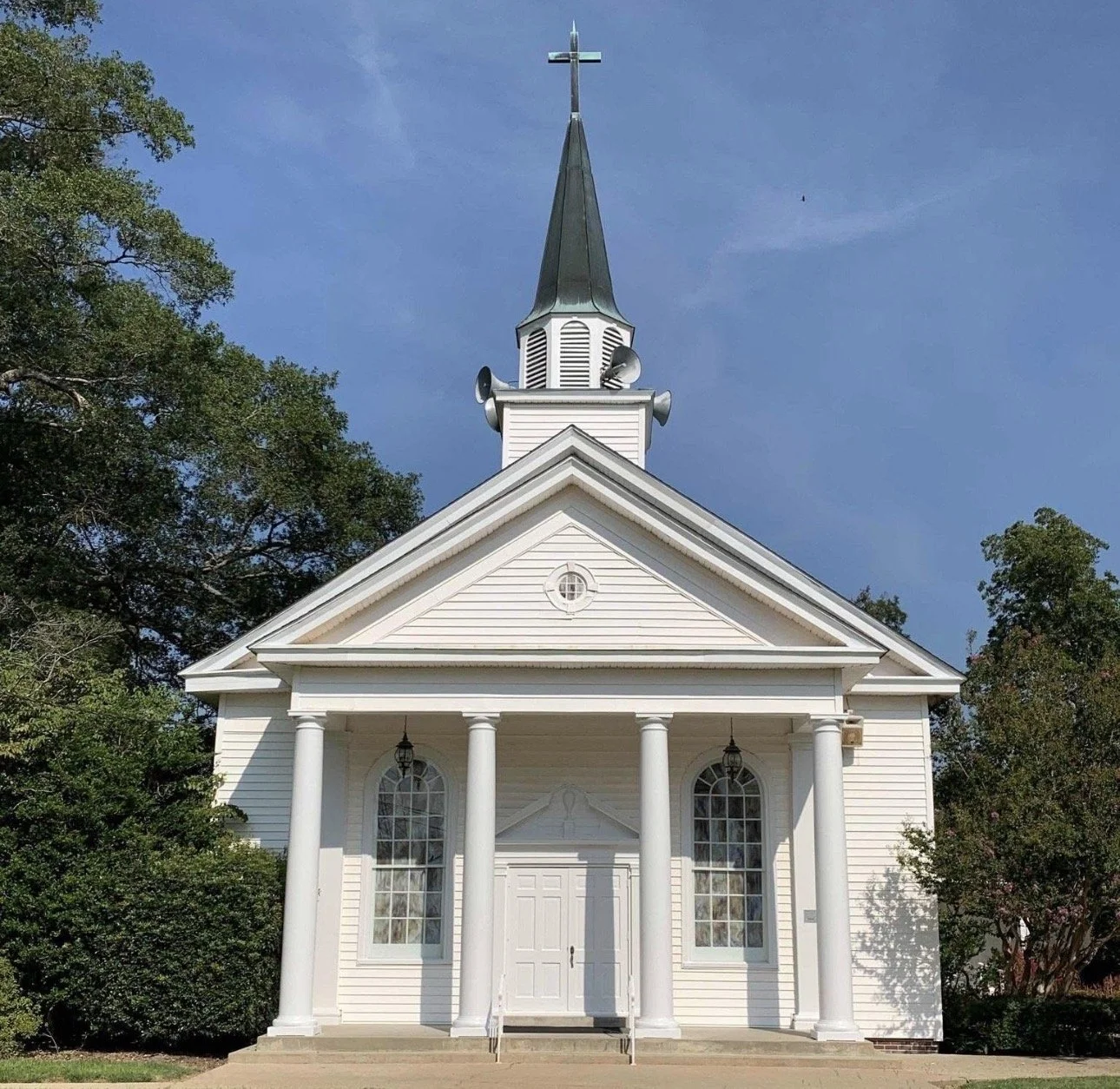 White church with tall steeple topped by a cross, surrounded by trees, under a blue sky.