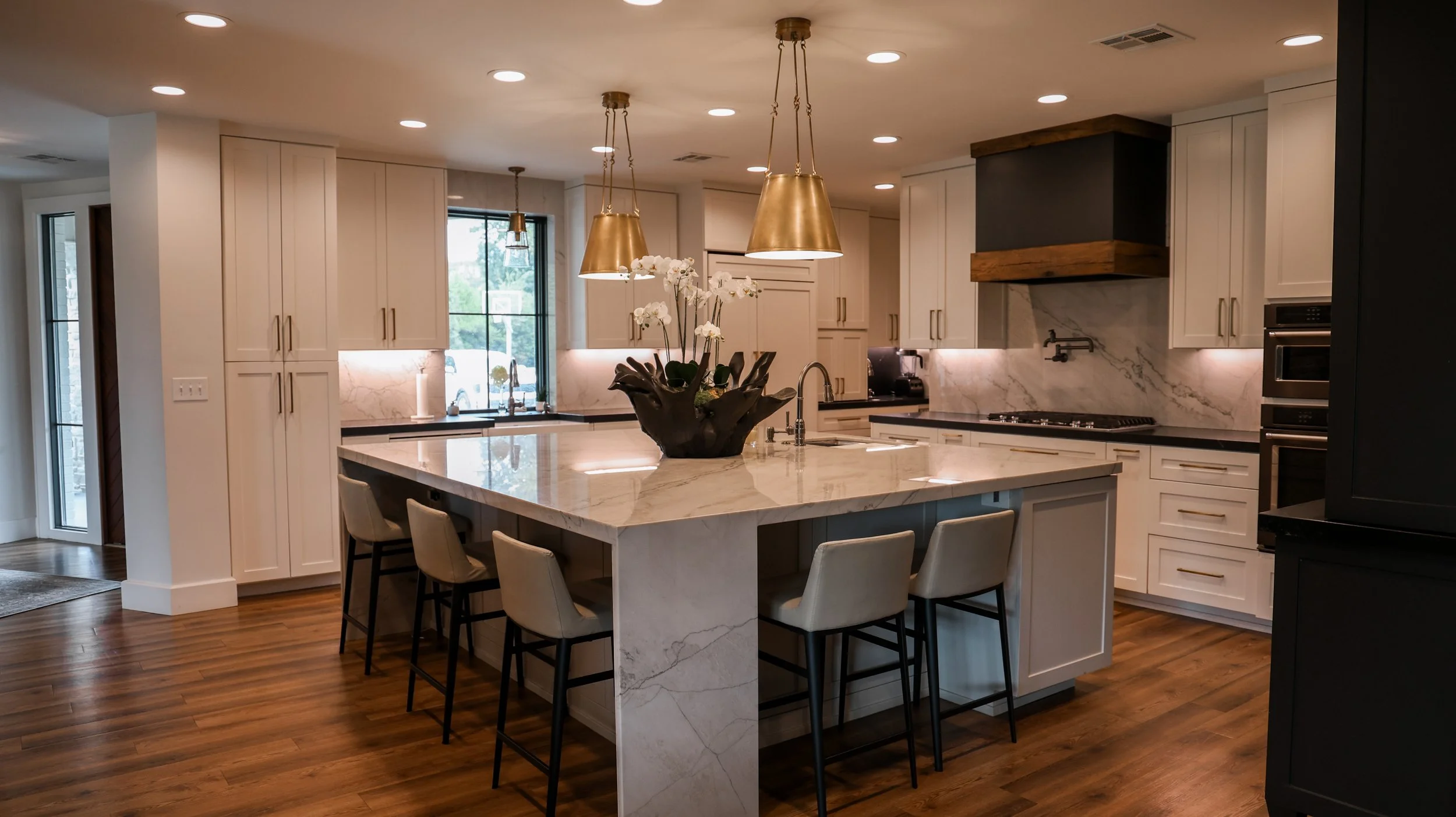 Modern kitchen with a large marble island, white cabinets, hardwood floors, gold pendant lights, and a flower arrangement on the island.