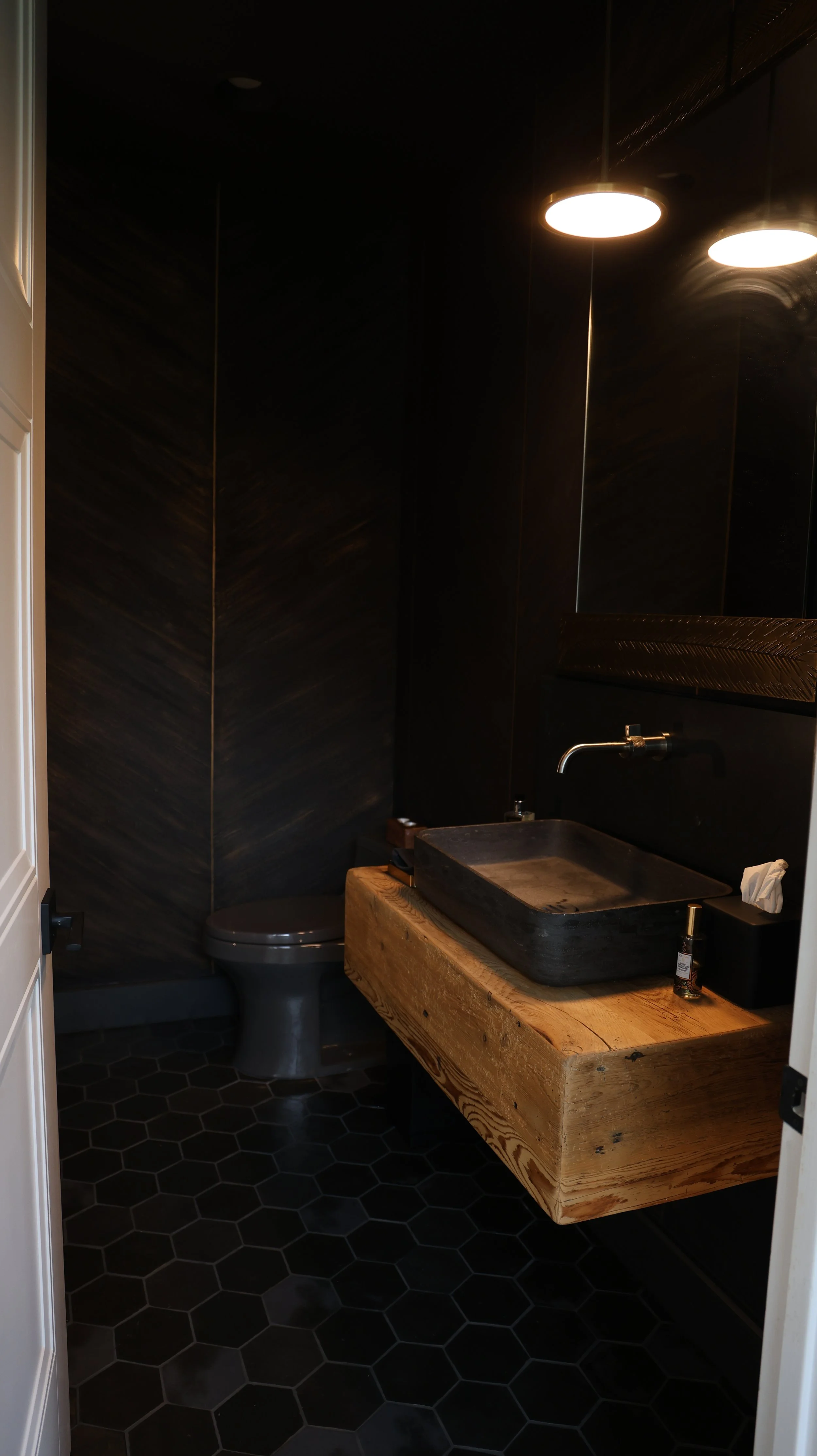 A small, dark bathroom with black walls, hexagonal black floor tiles, a black toilet, and a wooden vanity with a black stone sink and a modern faucet. Two ceiling lights illuminate the space.