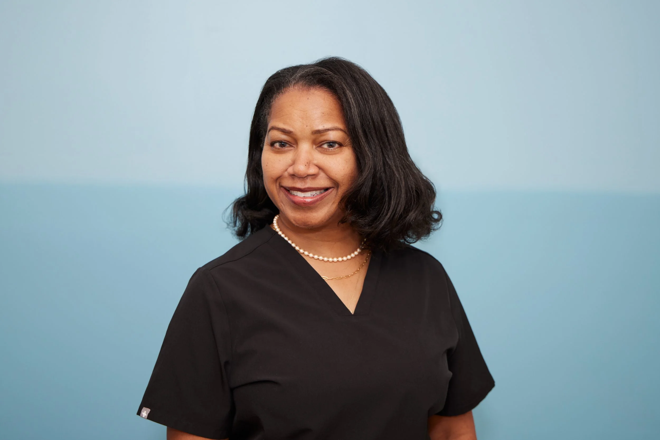 A smiling woman with shoulder-length black hair wearing black scrubs, a pearl necklace, and a gold chain necklace, standing against a two-tone blue background.