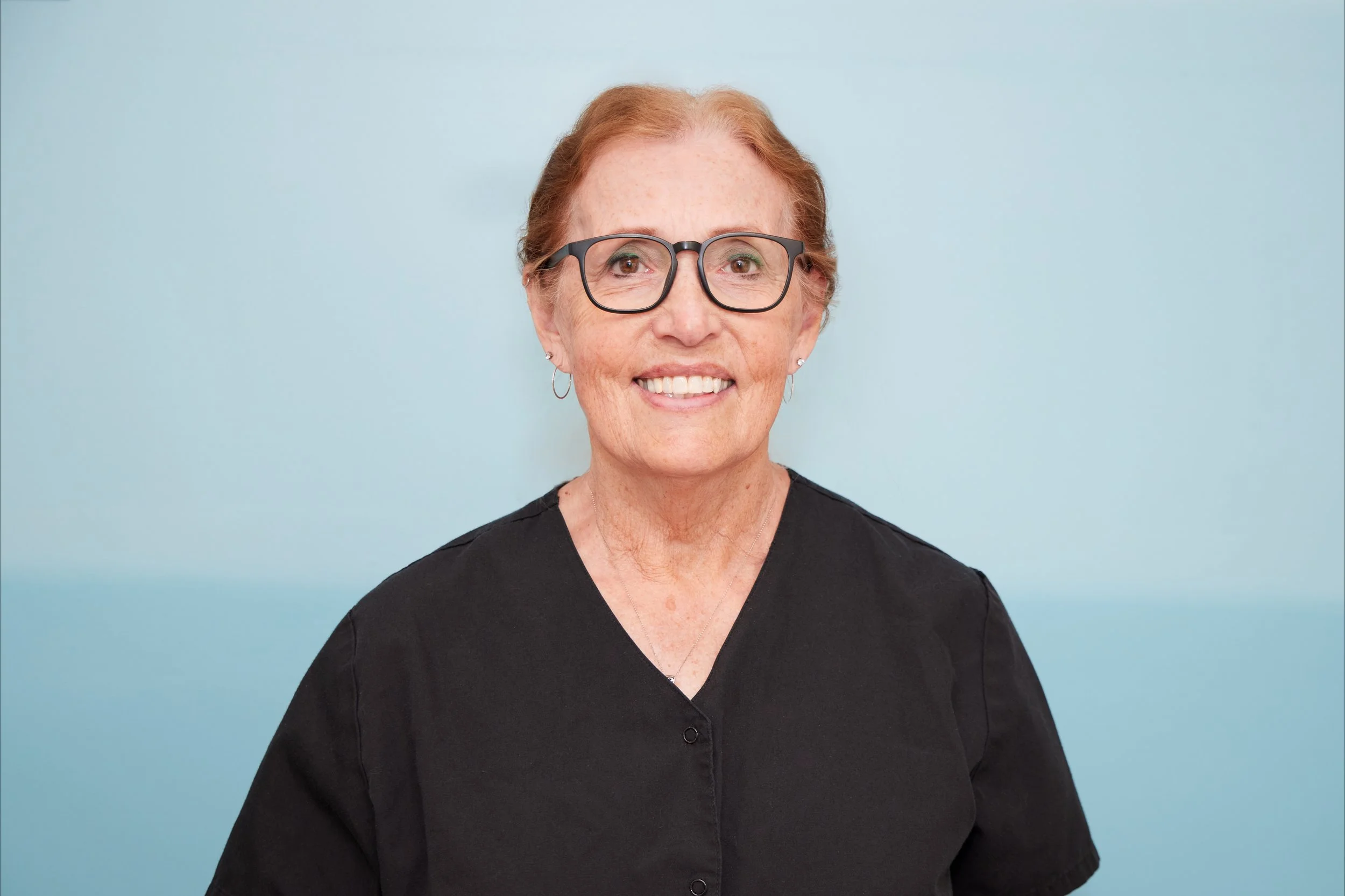 A smiling older woman with short red hair, wearing glasses and a black top, posing against a light blue background.
