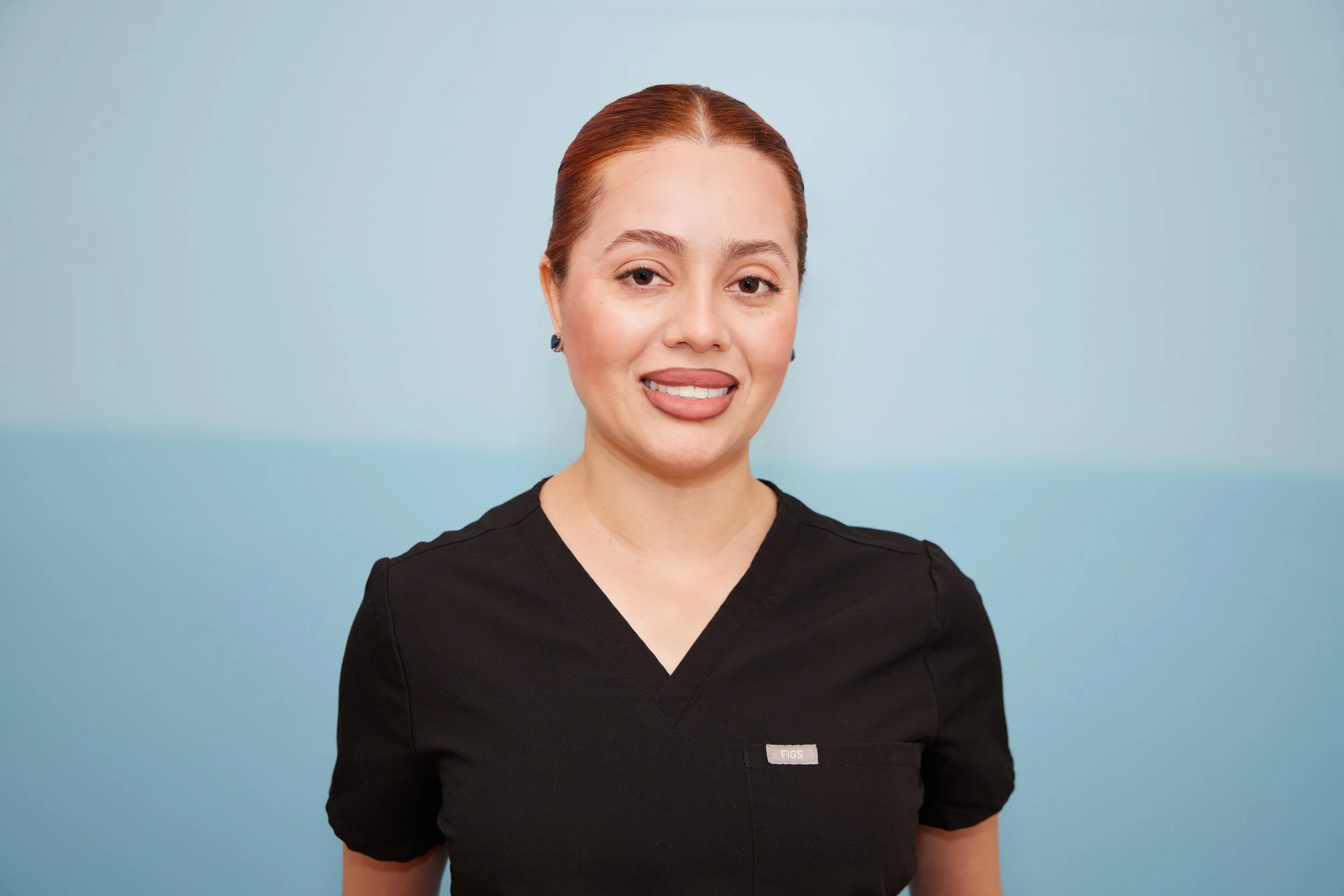A woman with red hair in a bun, wearing a black medical uniform with a name badge.