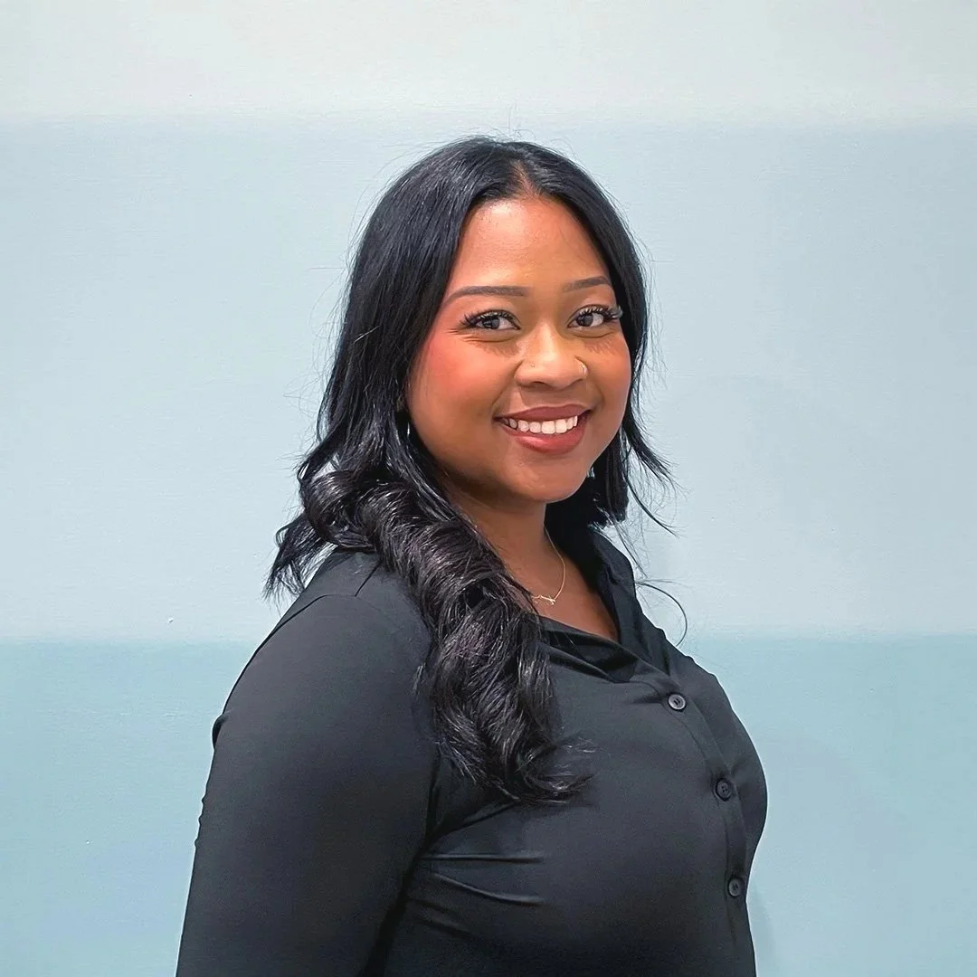 A woman with long, black, curled hair smiling and wearing a black button-up shirt against a plain light-colored wall.