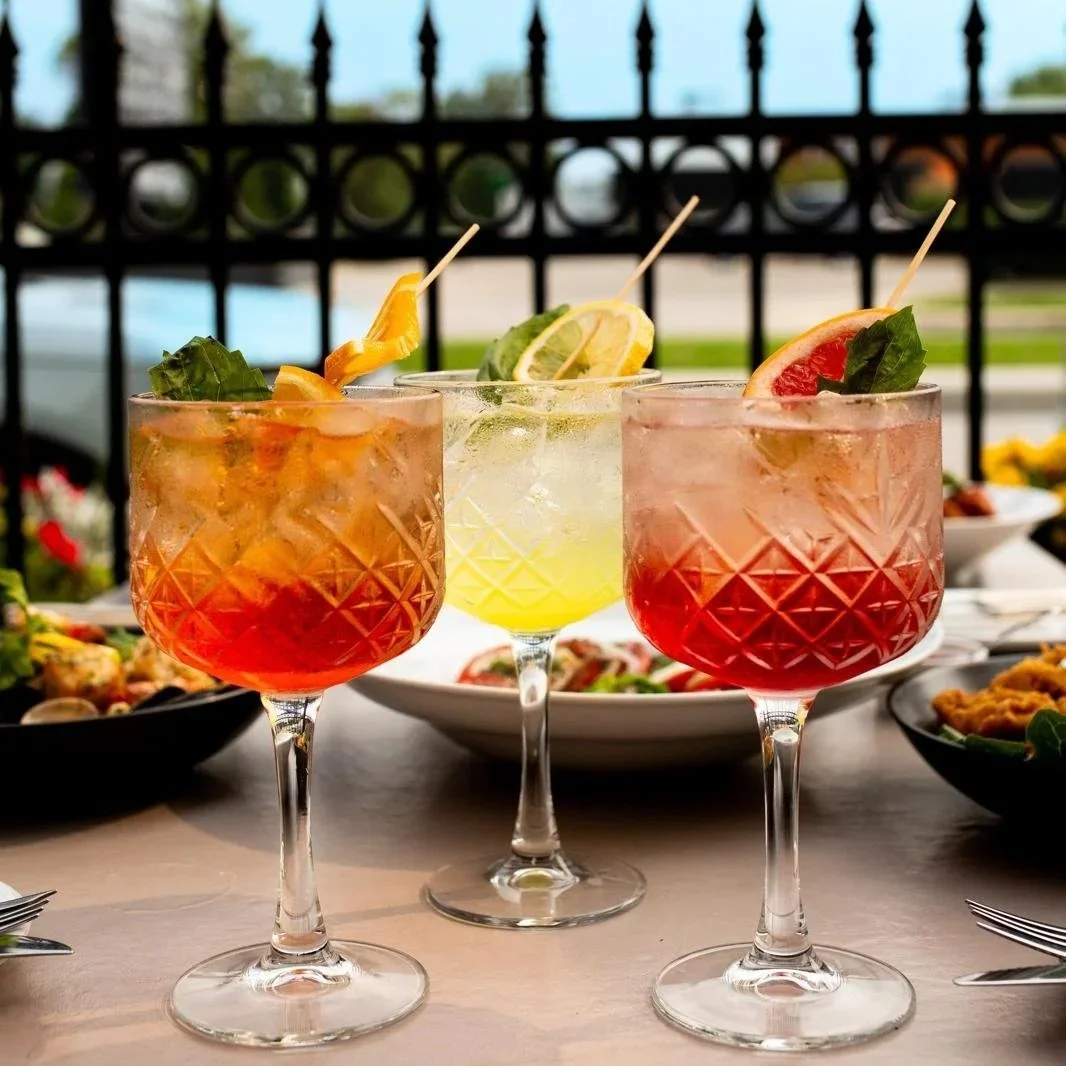 Three colorful cocktails with fruit garnishes on a table with food in the background.