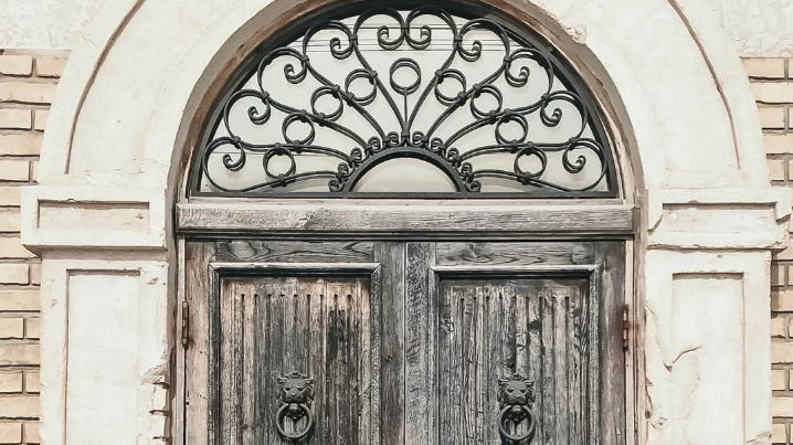 Old wooden door with metal lion head door knockers and intricate ironwork above it, set in stone and brick framework.