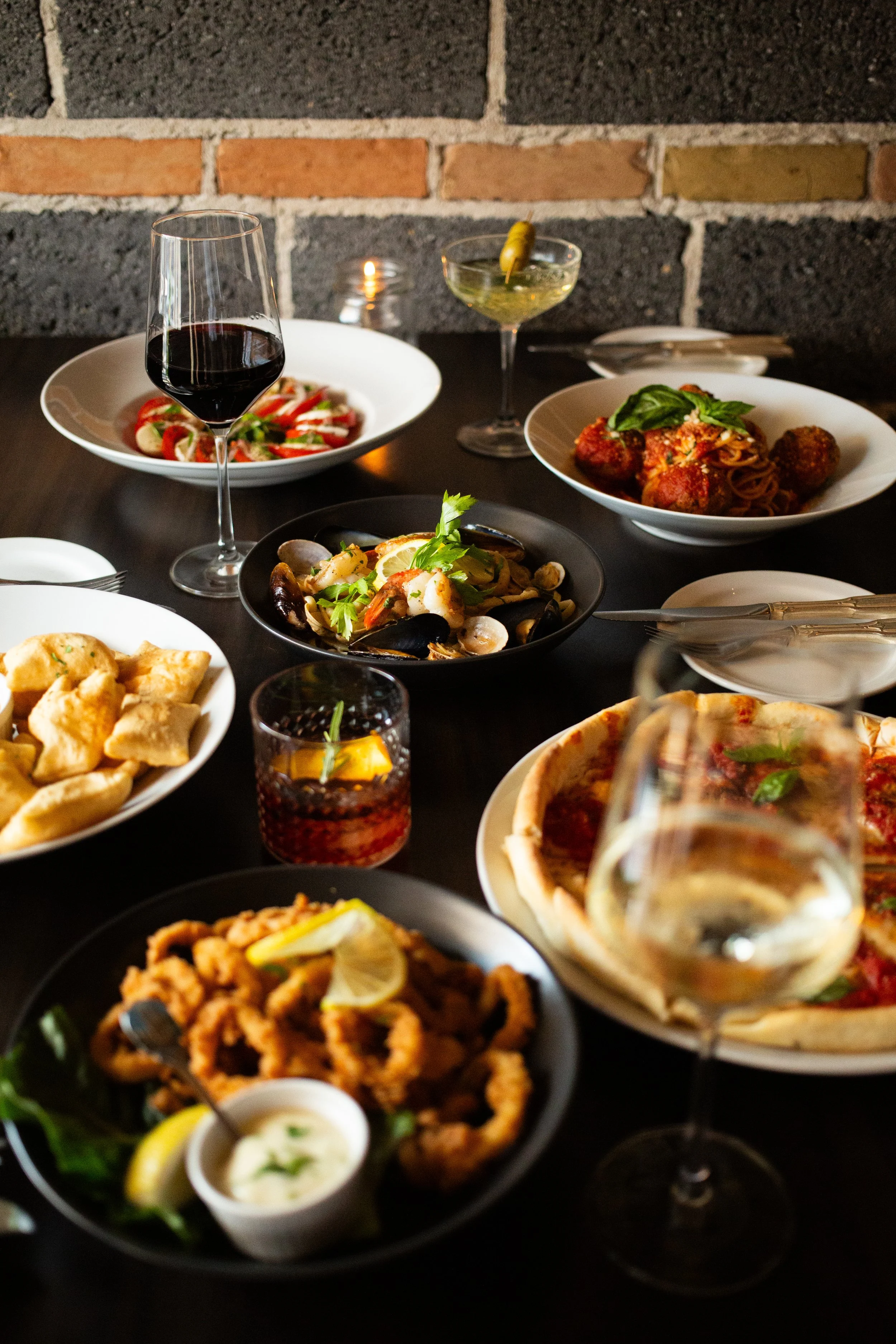 A table spread with various dishes including a pizza, fried calamari, pasta with meatballs, seafood, and salads, along with glasses of red and white wine and a cocktail, set against a black table and brick wall background.