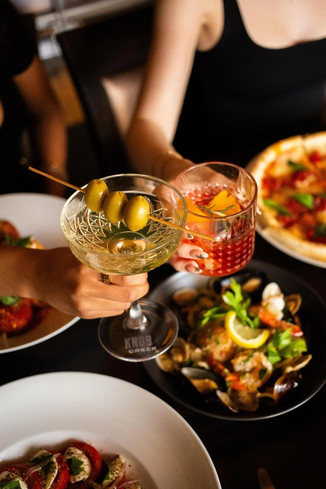 People clinking glasses at a dinner table with olives and a variety of food including pizza and seafood.