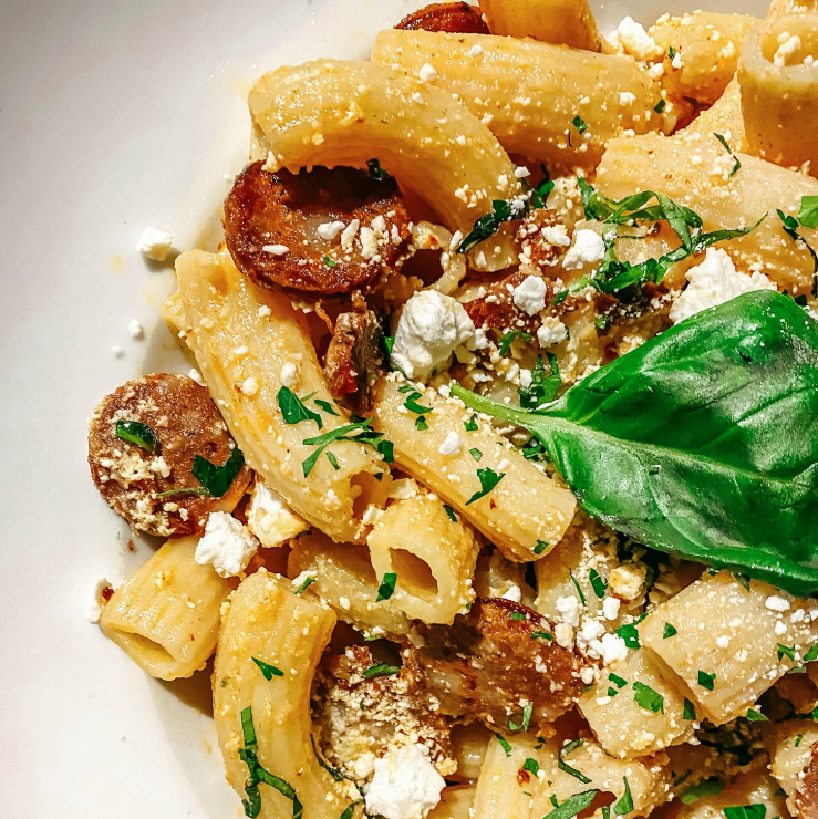 Close-up of cooked pasta with sausage slices, topped with crumbled cheese, chopped herbs, and a basil leaf on a white plate.