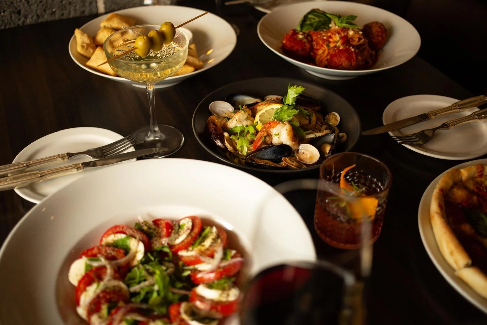 Family-style Italian meal on a dark table, including a seafood dish with shrimp, clams, and mussels garnished with herbs, a Caprese salad with tomato, mozzarella, and basil, a dish of meatballs with tomato sauce and basil, glasses of red sangria, a p