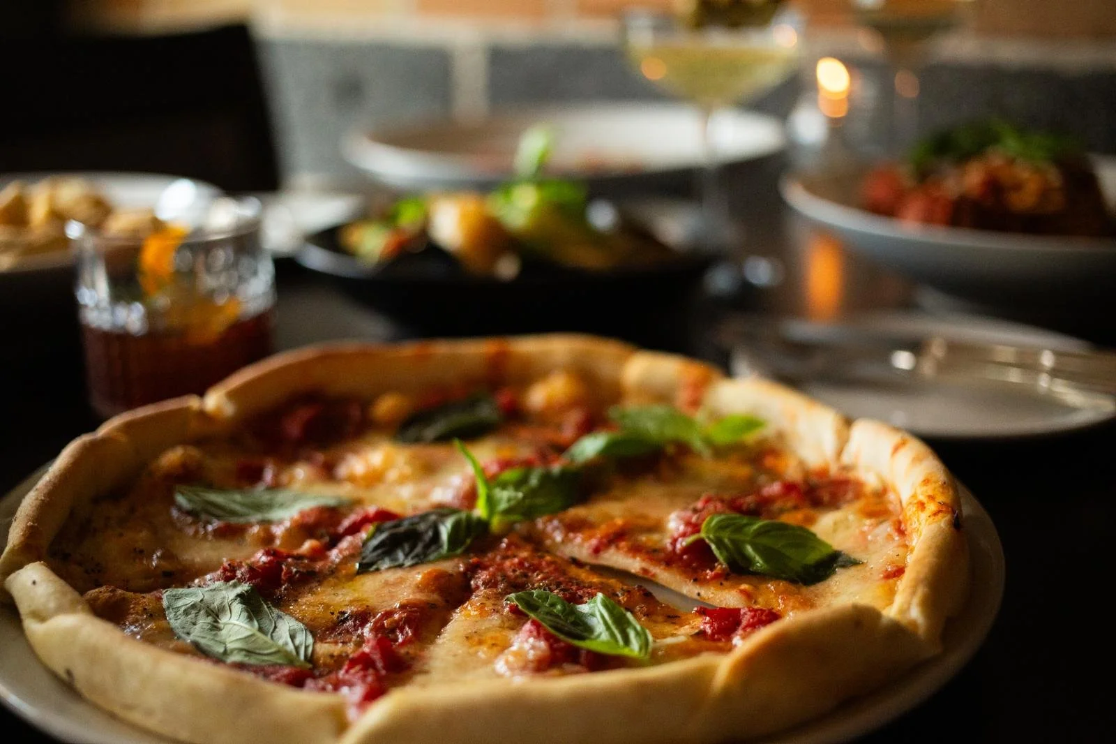 Close-up of a freshly baked margherita pizza with basil leaves on top, placed on a table with various dishes in the background.