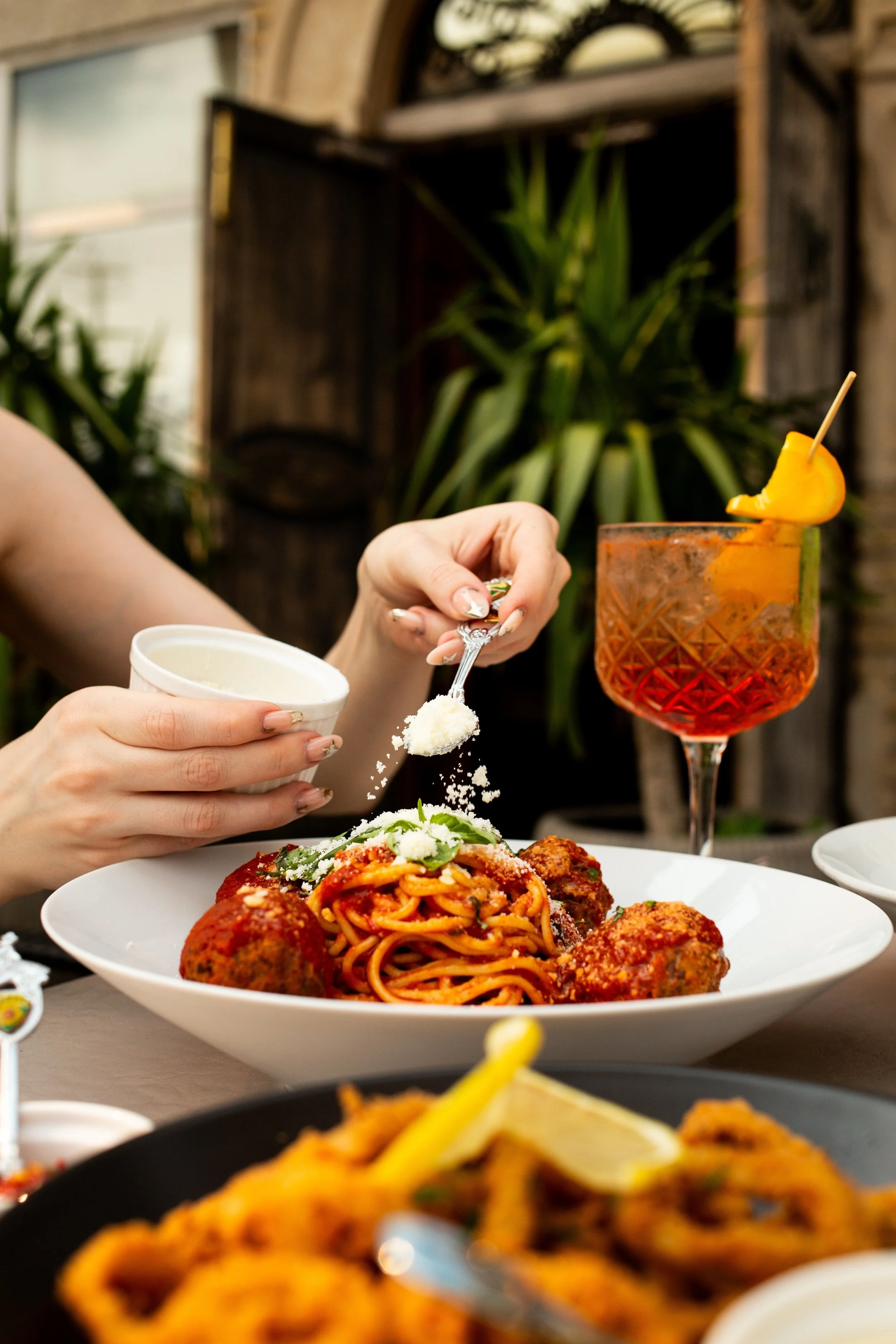 Person sprinkling grated cheese over a plate of spaghetti with meatballs, accompanied by a cocktail with an orange garnish.