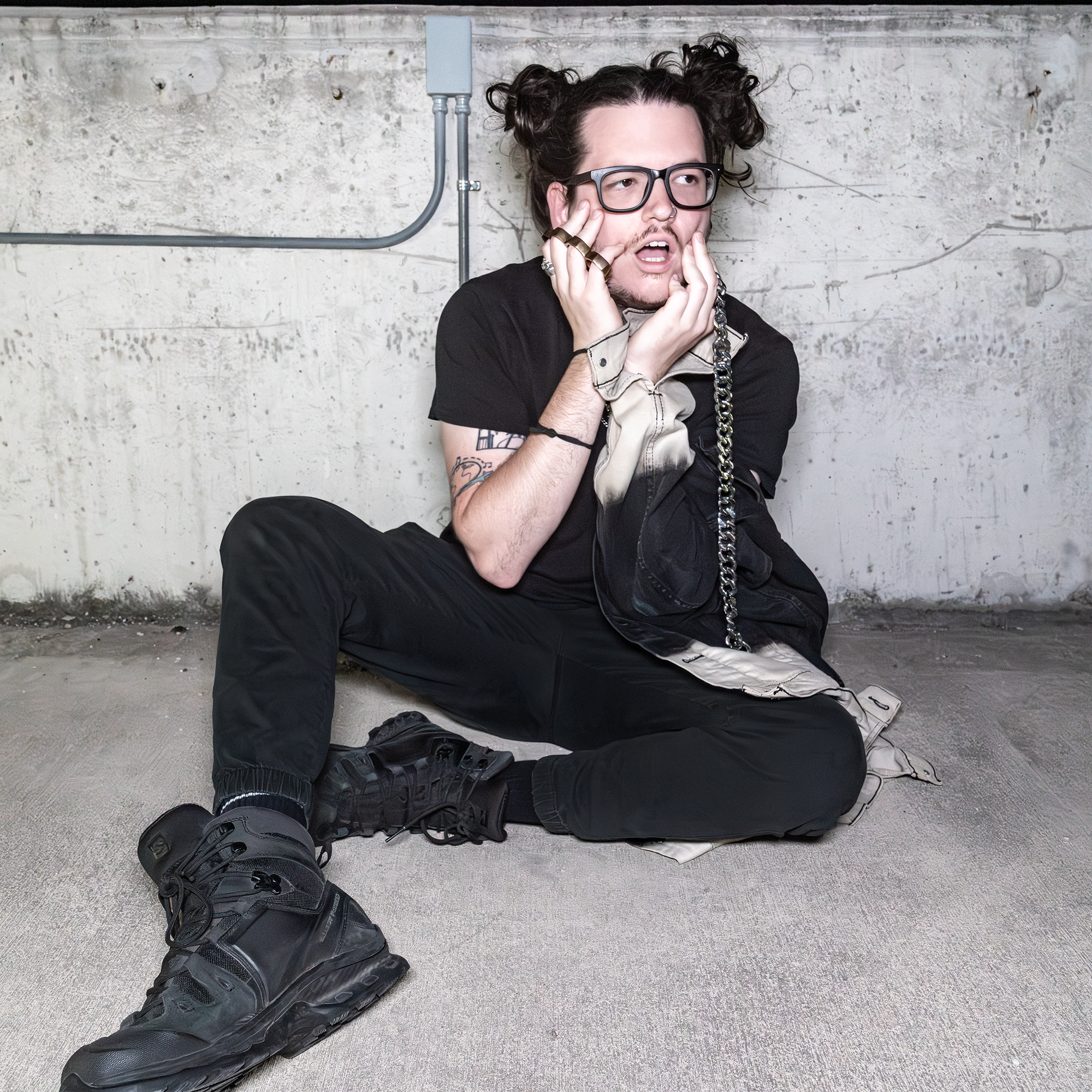 Person sitting on the floor in an industrial setting, wearing black clothes, chunky black boots, glasses, and strewn hair, holding face with a distressed expression.