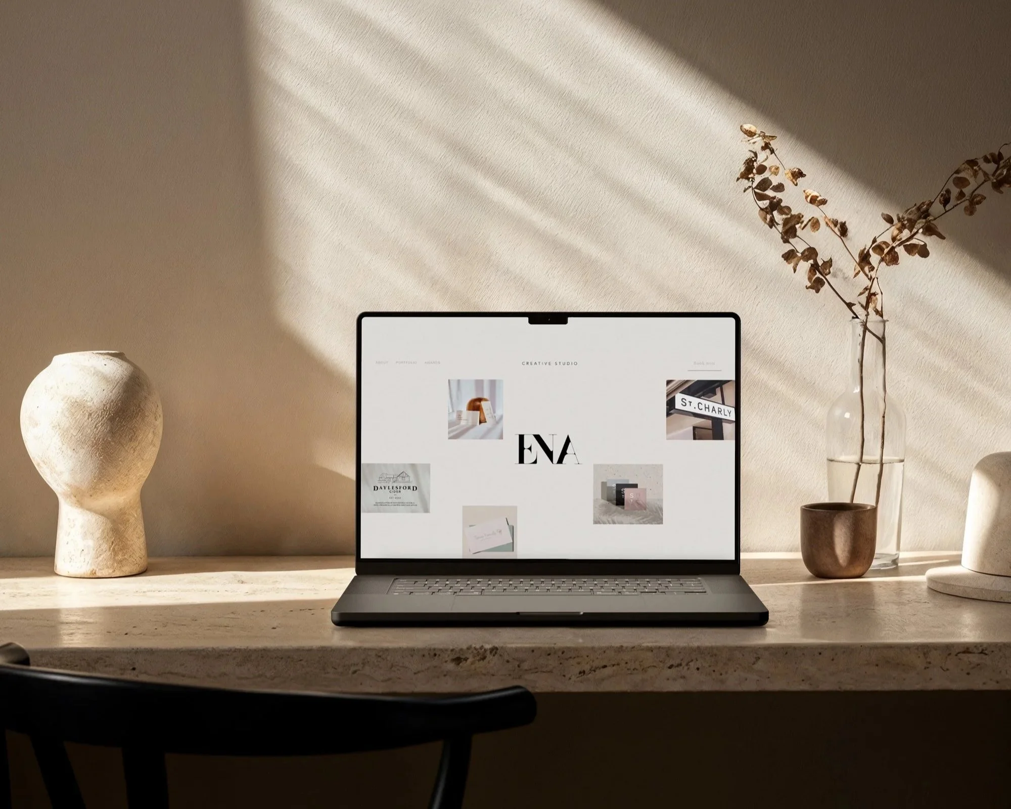 A laptop on a beige marble desk displaying a creative studio website, flanked by a textured white vase with a rounded top on the left and a glass vase with dried branches on the right, with sunlight creating shadows on a beige wall behind.
