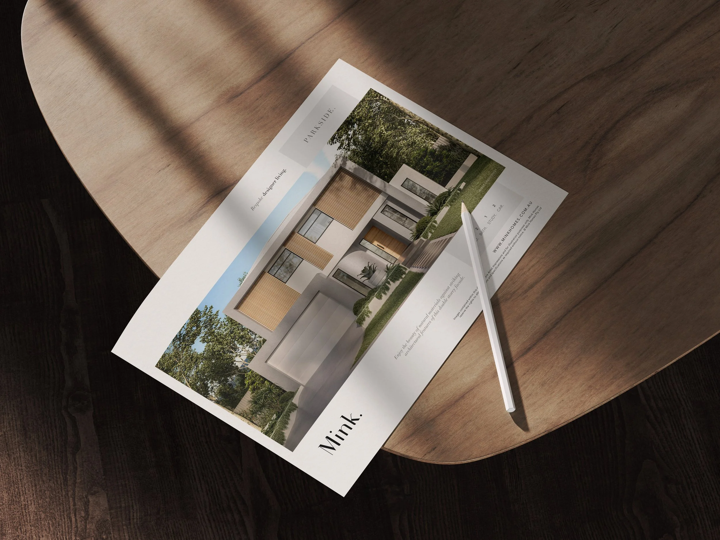 Brochure or flyer advertising a modern house design by Mink Homes, placed on a walnut wooden table with a white pen beside it.