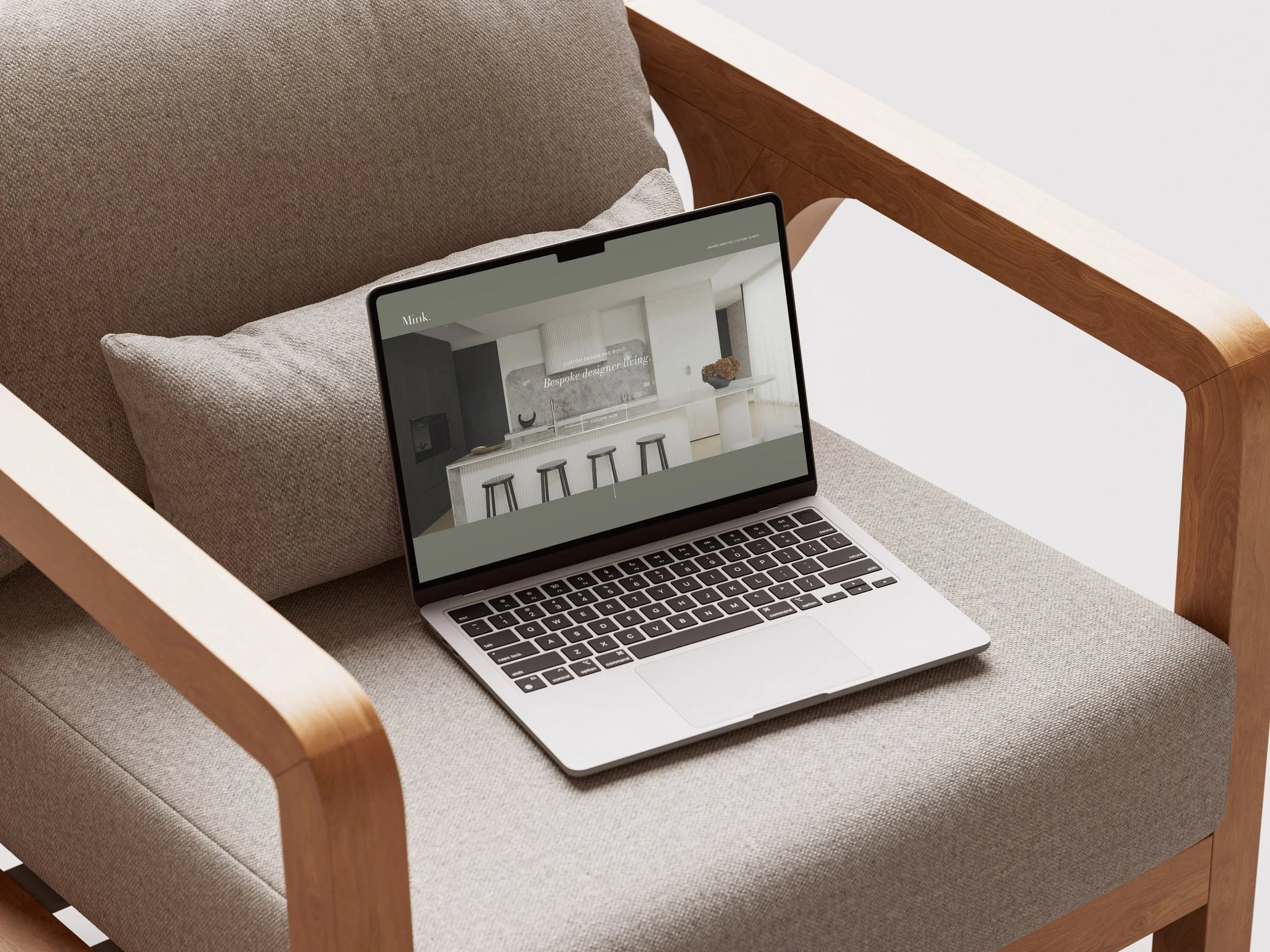 A laptop on a beige upholstered chair with wooden armrests, displaying a website for a bespoke custom home builder in Melbourne in a modern, minimalist kitchen.